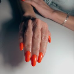 Gałczanka. Pędzlem i pęsetą - Zadbana dłoń z błyszczącymi, owalnymi paznokciami w intensywnym, koralowym odcieniu pomarańczy, podkreślonymi światłem. W tle druga dłoń z delikatną bransoletką.