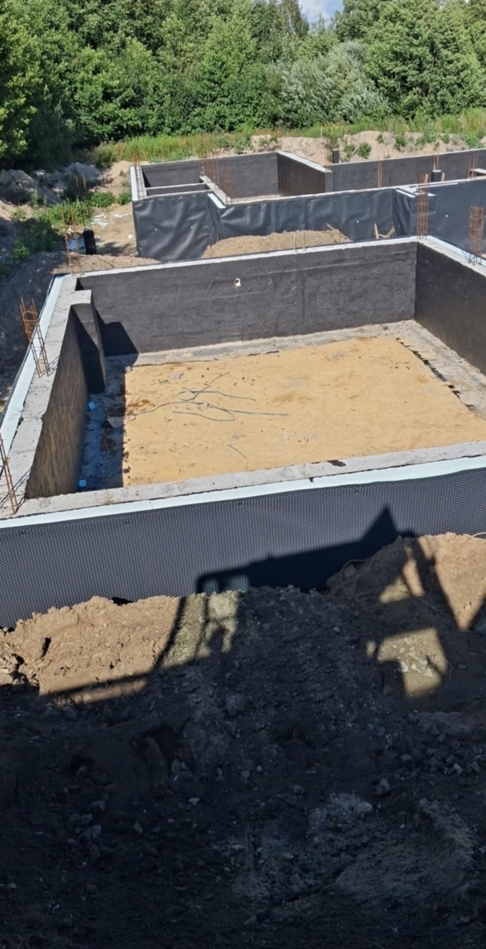 Wykopane i zabezpieczone fundamenty budynku mieszkalnego z widoczną czarną folią kubełkową, zbrojeniem i piaszczystym dnem. Ujęcie z góry, w tle zieleń.