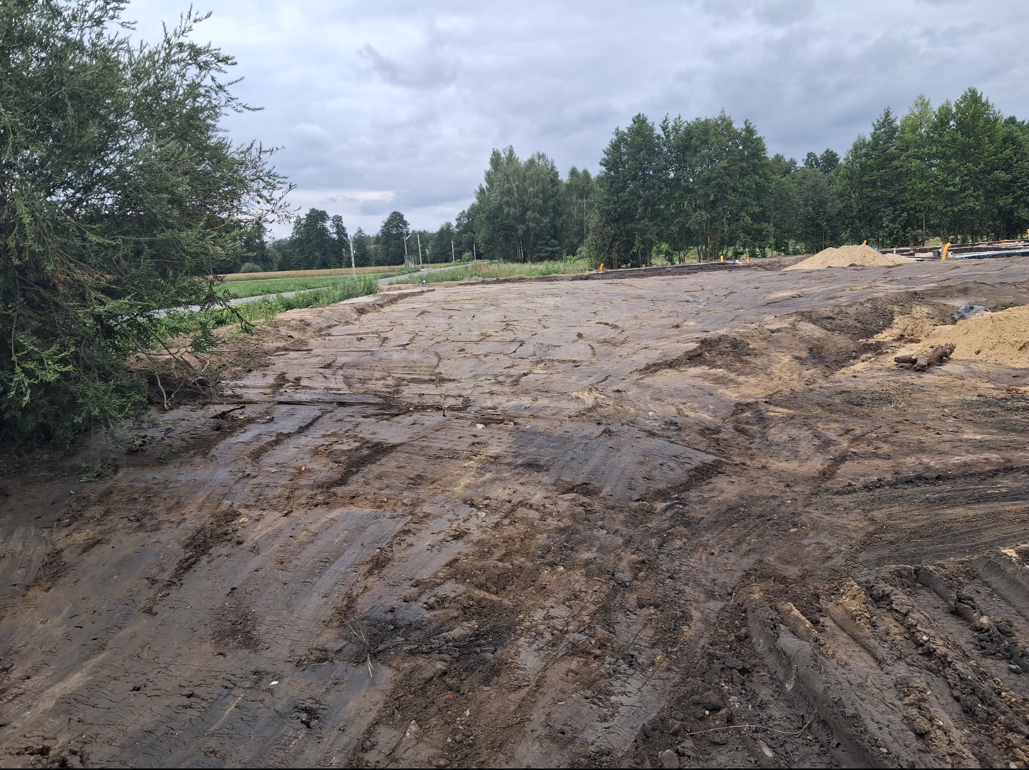 Rozległy plac budowy z ciemną, wilgotną ziemią pełną śladów maszyn, z widoczną hałdą jasnego piasku. W tle las i pochmurne niebo.