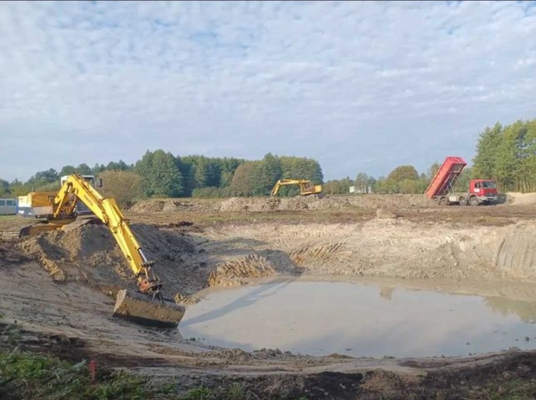 Żółta koparka pogłębia rozległy wykop z błotnistą wodą, obok czerwona wywrotka i druga koparka na placu robót ziemnych.