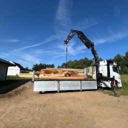 WANGER POLSKA Piotr Kosman - Biała ciężarówka z żurawiem HDS FASSI F800XP na otwartym terenie, z ładunkiem jasnych desek budowlanych, gotowa do rozładunku przy nowo budowanych domach.