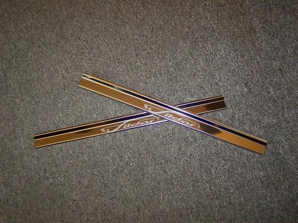 Dwie błyszczące, metalowe nakładki progowe z napisem 'Sildar' ułożone na szarym, teksturowanym tle.