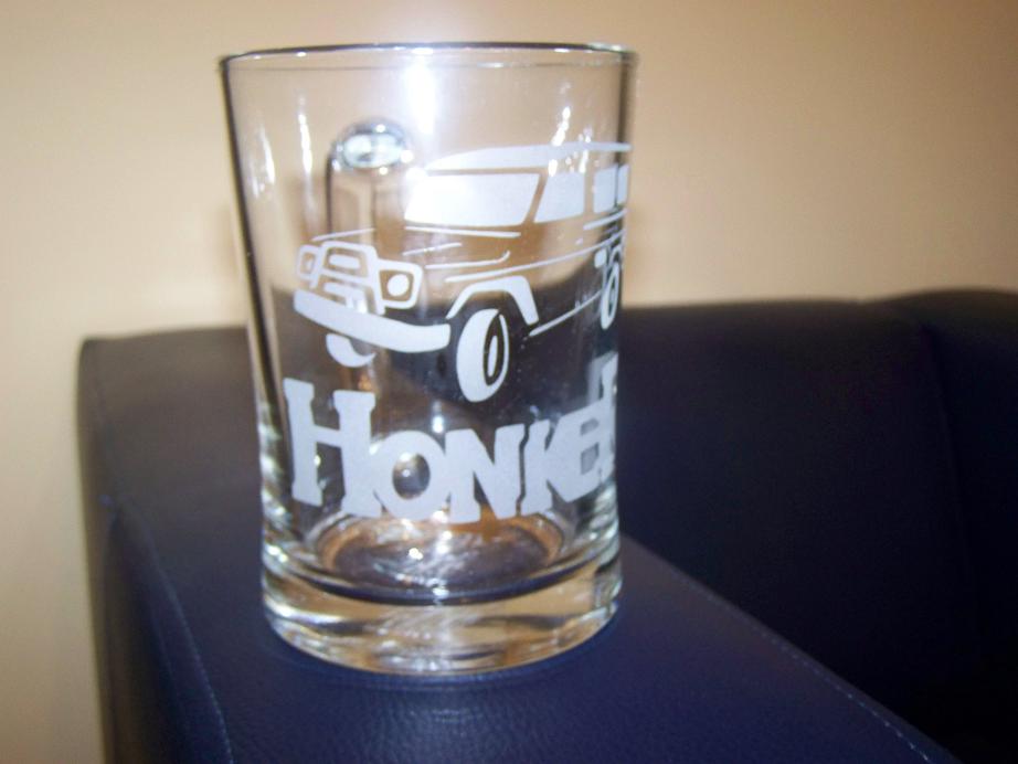 Szklanka z grawerem przedstawiającym pojazd typu kamper i napis 'Honker', stojąca na ciemnym, skórzanym fotelu.