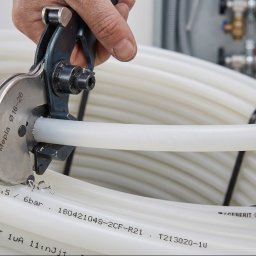 Domgard - Instalacje Optymalne - Ręka instalatora precyzyjnie tnąca białą rurę PEX/PERT ze zwoju specjalnymi nożycami Mepla Ø16-26, z elementami rozdzielacza instalacji w tle.