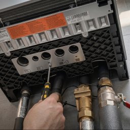 Domgard - Instalacje Optymalne - Ręka technika dokręcająca śrubę w dolnej części kotła gazowego Viessmann Vitodens 200-G, z widocznymi rurami, zaworami i izolacją na tle szarych płytek.