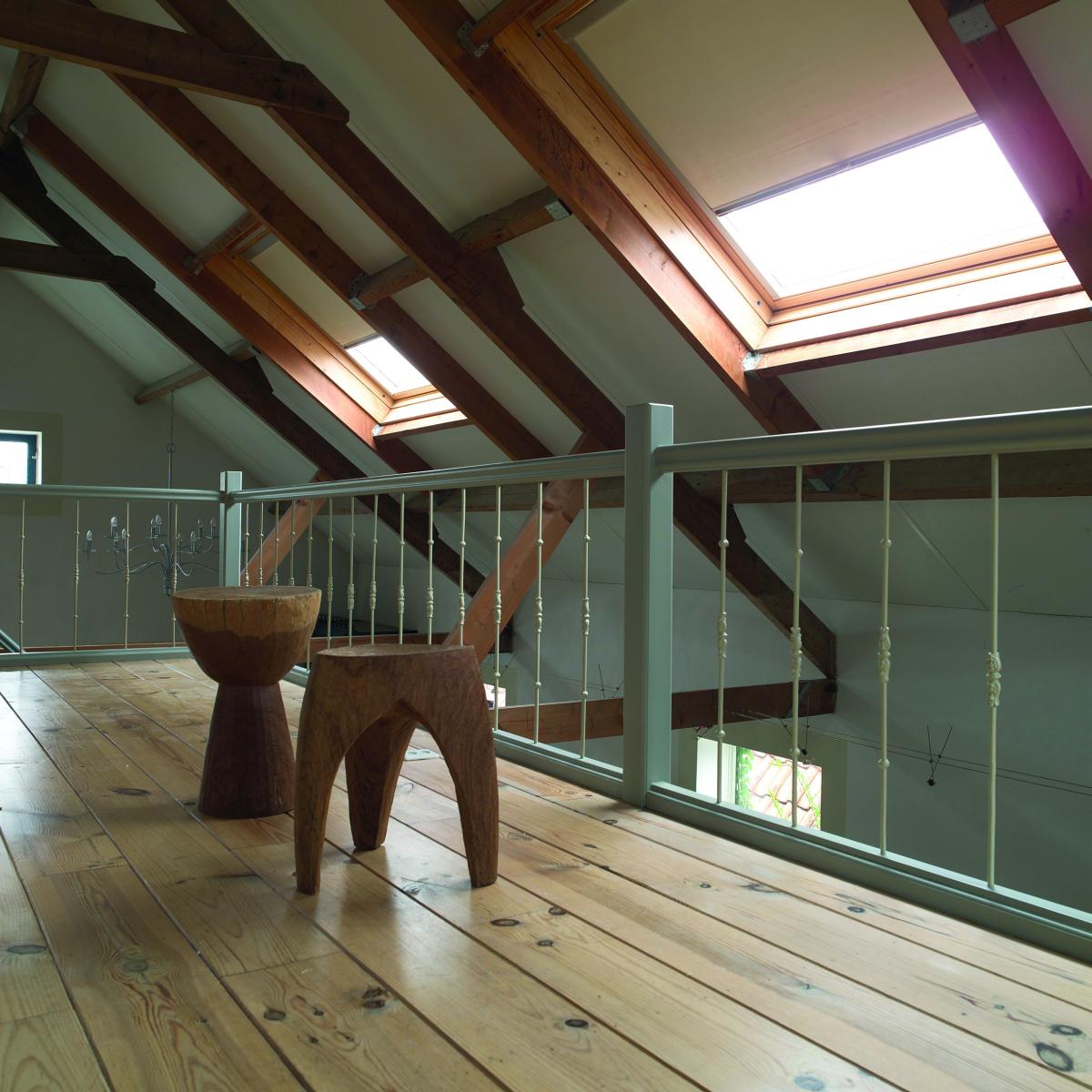 Poddasze z drewnianą podłogą i balustradą, widoczne drewniane krokwie i okna dachowe z zasłoniętymi roletami, dwa drewniane stołki na pierwszym planie.