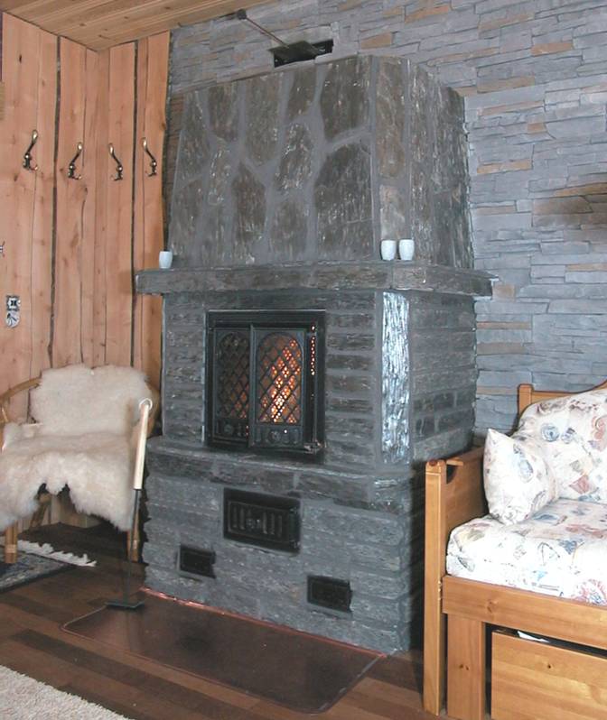 Kamienny piec z żeliwnymi drzwiczkami, widoczne płomienie w palenisku, rustykalne wnętrze z drewnianą boazerią i kamienną ścianą.