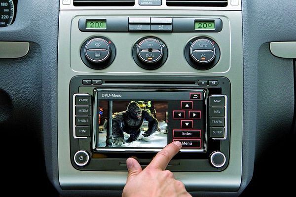 Wnętrze samochodu z systemem nawigacji RNS 510 wyświetlającym film, palec dotyka ekranu, widoczne przyciski sterowania klimatyzacją i nawigacją.