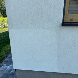 AquaBruk - Fragment jasnej elewacji budynku z wyraźną linią podziału między czystą a zabrudzoną powierzchnią, obok okna z drewnianą ramą. Na dole kostka brukowa z resztkami piany po myciu.