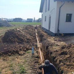 EKO-BUD USŁUGI WODNO-KANALIZACYJNE, BUDOWA PRZYŁĄCZY - Pracownik układa pomarańczowe rury w głębokim wykopie obok nowo wybudowanego białego domu z ciemnym dachem, na tle otwartego terenu.