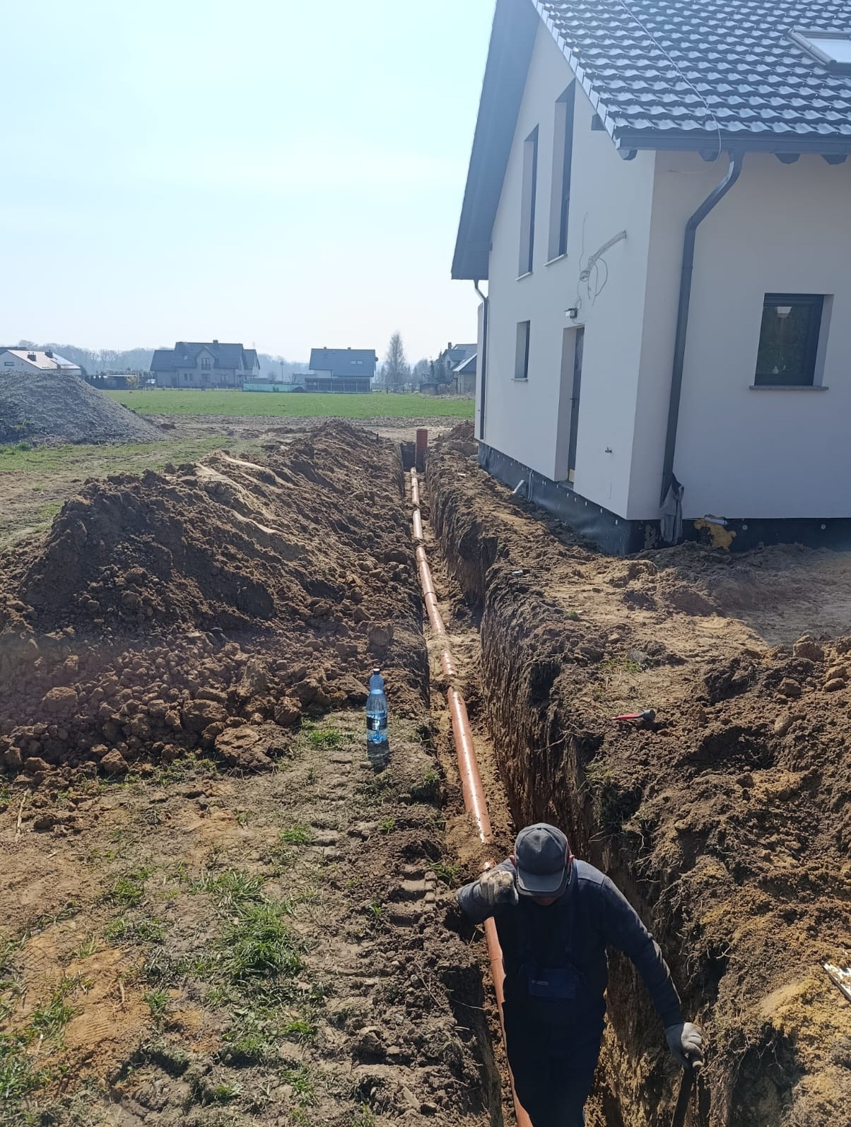 Pracownik układa pomarańczowe rury w głębokim wykopie obok nowo wybudowanego białego domu z ciemnym dachem, na tle otwartego terenu.