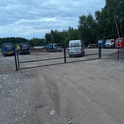 KBS profil - Czarna, dwuskrzydłowa brama panelowa z siatki, otwierająca wjazd na plac transportowy z ciężarówkami i busem KBS PROFIL, pod zachmurzonym niebem.