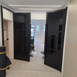 JTM SPÓŁKA Z OGRANICZONĄ ODPOWIEDZIALNOŚCIĄ - Eleganckie, dwuskrzydłowe drzwi wewnętrzne z czarnymi, metalowymi profilami i ciemnym szkłem, zamontowane w jasnym korytarzu z podłużnym oświetleniem LED, obok widoczne narzędzia.