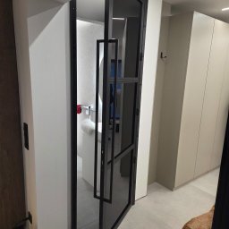 JTM SPÓŁKA Z OGRANICZONĄ ODPOWIEDZIALNOŚCIĄ - Nowoczesne, czarne drzwi wewnętrzne z wąskimi profilami, podziałami szklanymi i podłużnym uchwytem, otwarte na jasne wnętrze z szafami, na podłodze pies.