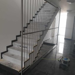 JTM SPÓŁKA Z OGRANICZONĄ ODPOWIEDZIALNOŚCIĄ - Nowoczesna klatka schodowa z pionowymi, metalowymi słupkami i poziomym uchwytem, schody zabezpieczone folią ochronną, na błyszczącej, ciemnej posadzce leży przedłużacz.