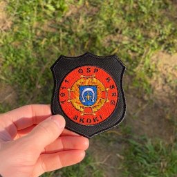 Haftosław - Haftowana naszywka w kształcie tarczy z logo OSP Skoki, z czerwonym tłem i złotymi detalami, trzymana w dłoni na tle rozmytej zielonej trawy.