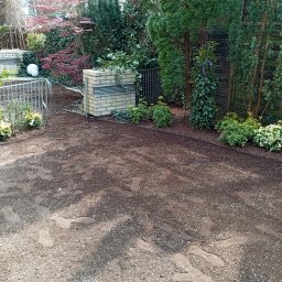 GARDENPROMAX S.P. Z.O.O - Przygotowany grunt w ogrodzie z widoczną mieszanką ziemi i jasnych granulek, otoczony zielonymi krzewami, czerwonym klonem i iglakami. W tle wąż ogrodowy i murki z cegły.