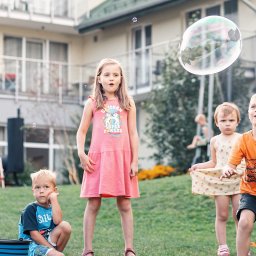 Holimo Hotel Restauracja & SPA - Grupa dzieci z otwartymi ustami i zachwytem obserwuje ogromną, opalizującą bańkę mydlaną unoszącą się nad zielonym trawnikiem, w tle budynek.