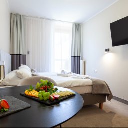Holimo Hotel Restauracja & SPA - Komfortowy pokój z dużym łóżkiem, telewizorem na ścianie i okrągłym stolikiem. Na nim taca z owocami, serami, dwoma kieliszkami szampana i kartą powitalną. Jasne zasłony w tle.