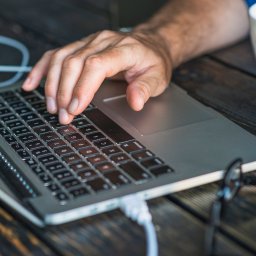 DG Networks - Męska dłoń na klawiaturze i trackpadzie srebrnego laptopa, podłączonego białym kablem, leżącego na ciemnym, postarzanym drewnianym blacie. W tle biały kubek i okulary.