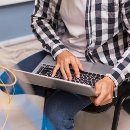 DG Networks - Mężczyzna w koszuli w kratę pracuje na laptopie, konfigurując system w serwerowni, z widocznymi żółtymi kablami sieciowymi i częściowo uciętym regałem.