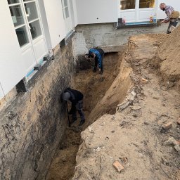 TOMBUD - Trzech pracowników ręcznie pogłębia wykop wzdłuż ściany białego budynku z dużymi oknami, odsłaniając fundamenty i przygotowując teren pod dalsze prace.