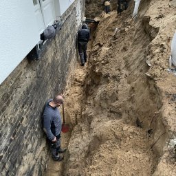 TOMBUD - Trzech pracowników w głębokim wykopie przy fundamencie domu z białym tynkiem, jeden oparty o ceglaną ścianę, inni kopią ziemię wokół ciemnego zbiornika.