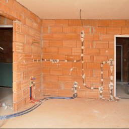 MC Electro Service - Rozprowadzone instalacje elektryczne i wodne na ścianach z czerwonych pustaków ceramicznych w pomieszczeniu w stanie deweloperskim, z widocznymi peszlami i puszkami.