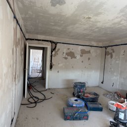 MC Electro Service - Pokój w trakcie remontu z poprowadzonymi po ścianach i suficie czarnymi i niebieskimi peszlami instalacji elektrycznej. Na podłodze narzędzia Bosch i zwinięte przewody.