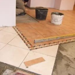 Układanie płytek podłogowych z dekoracyjnym pasem mozaiki, widoczne dwa wiadra i poziomica, w tle pracownik w trakcie układania płytek.