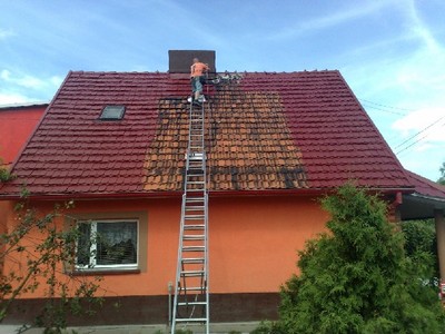 Pracownik na drabinie maluje dach z czerwonej dachówki, widoczna różnica koloru między pomalowaną a niepomalowaną częścią dachu, pomarańczowa elewacja budynku.