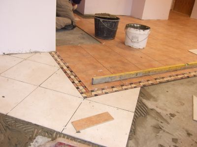 Układanie płytek podłogowych z dekoracyjnym pasem mozaiki, widoczne dwa wiadra i poziomica, w tle pracownik w trakcie układania płytek.