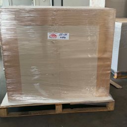 Inter-Papier - Duża paleta z arkuszami papieru lub tektury, szczelnie owinięta folią stretch i taśmą, z etykietą 'INTERPAPIER' (55x35, 418 kg), stojąca na betonowej posadzce magazynu.
