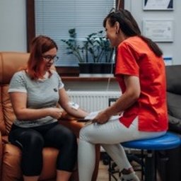 Centrum medyczne VitMeUp Wrocław - Kobieta w okularach otrzymująca kroplówkę dożylną w brązowym fotelu, obok uśmiechnięta osoba w czerwonej koszulce monitorująca zabieg. W tle okno z żaluzjami i certyfikaty na ścianie.