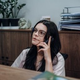 Centrum medyczne VitMeUp Wrocław - Młoda kobieta w okularach z ciemnymi włosami, zamyślona, rozmawia przez telefon stacjonarny przy drewnianym biurku. W tle roślina i dokumenty.