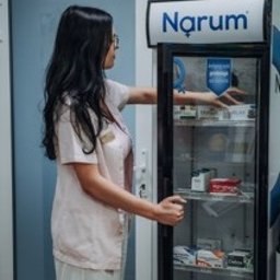 Centrum medyczne VitMeUp Wrocław - Kobieta w jasnym uniformie medycznym otwiera przeszkloną, wąską lodówkę z logo 'Narum', sięgając po produkt z półki. W tle jasne ściany.