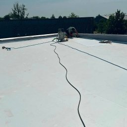 BIST BUILDERS - Specjalista w zielonej koszulce pracuje na płaskim dachu, aplikując jasną powłokę izolacyjną, z widocznym sprzętem i długimi wężami.
