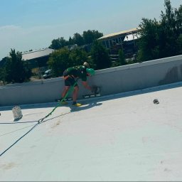 BIST BUILDERS - Pracownik w zielonej koszulce i uprzęży bezpieczeństwa wykonuje prace wykończeniowe na krawędzi płaskiego dachu, z widocznymi zrolowanymi rurami i słonecznym niebem.