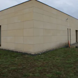 Elewacja budynku wykończona jasnymi, kwadratowymi płytami kamiennymi, widoczne okna i komin, trawnik z przodu, w tle budynek z cegły.