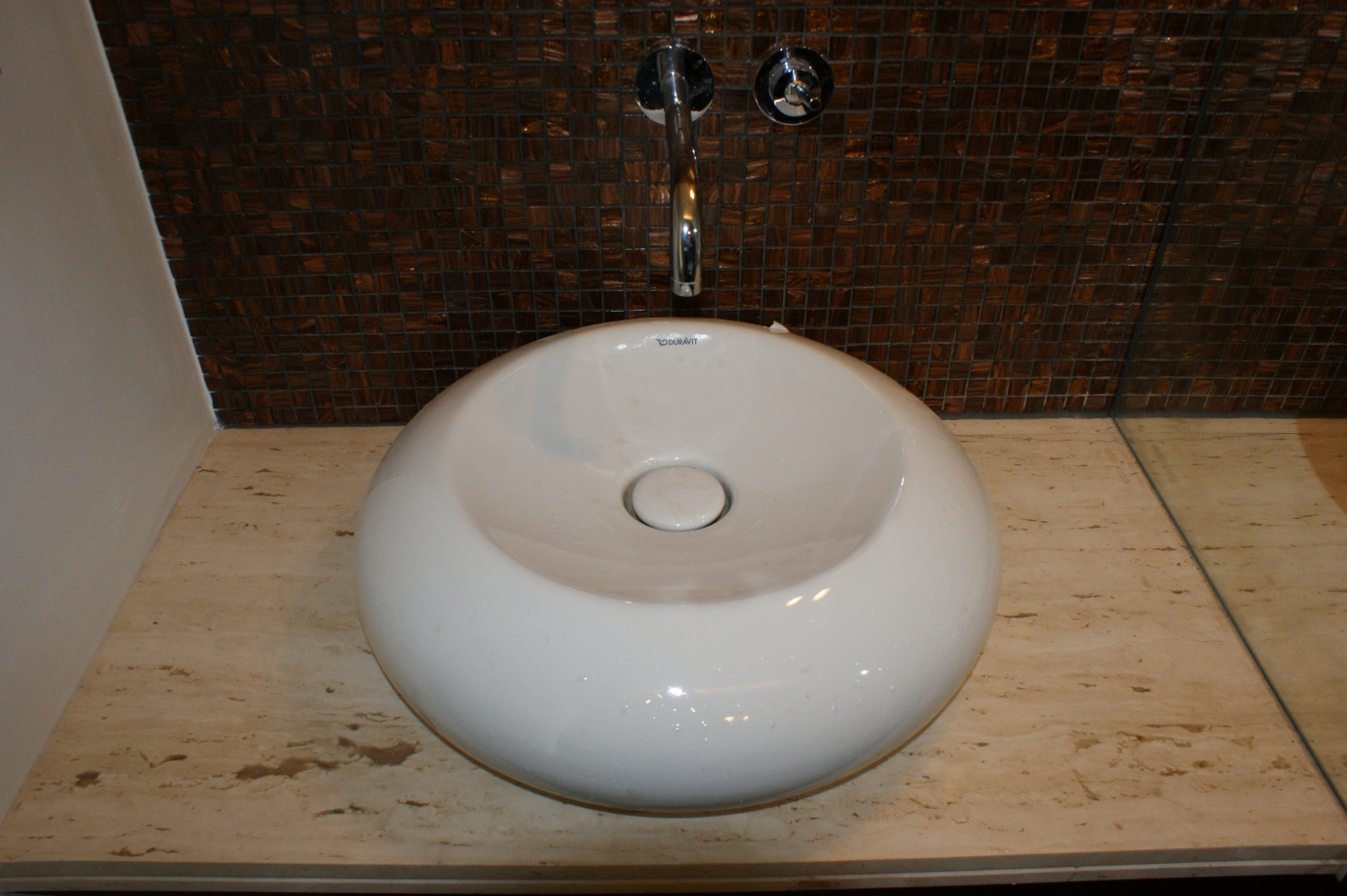 Biała, okrągła umywalka nablatowa marki Duravit na blacie z trawertynu, z mozaiką w odcieniach brązu na ścianie w tle i chromowaną armaturą.