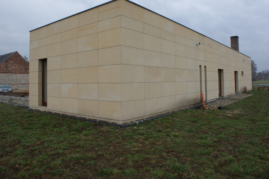 Elewacja budynku wykończona jasnymi, kwadratowymi płytami kamiennymi, widoczne okna i komin, trawnik z przodu, w tle budynek z cegły.