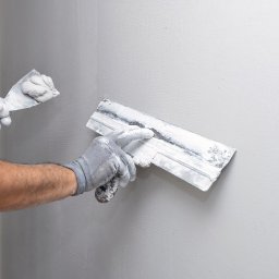Home Serwiss - Ręce w szarych rękawicach nakładają świeżą gładź na szarą ścianę za pomocą szerokiej szpachli, obok mniejsza szpachelka z zapasem masy.