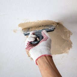 Home Serwiss - Ręka w białej rękawicy z czerwonymi kropkami precyzyjnie nakłada masę wykończeniową szpachlą na białą, chropowatą powierzchnię, przygotowując ją do dalszych prac.