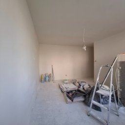 Home Serwiss - Pokój w trakcie remontu z gładkimi, jasnymi ścianami i skośnym sufitem. Na podłodze worki z materiałami, wiadra z farbą i drabina, wskazujące na prace wykończeniowe.