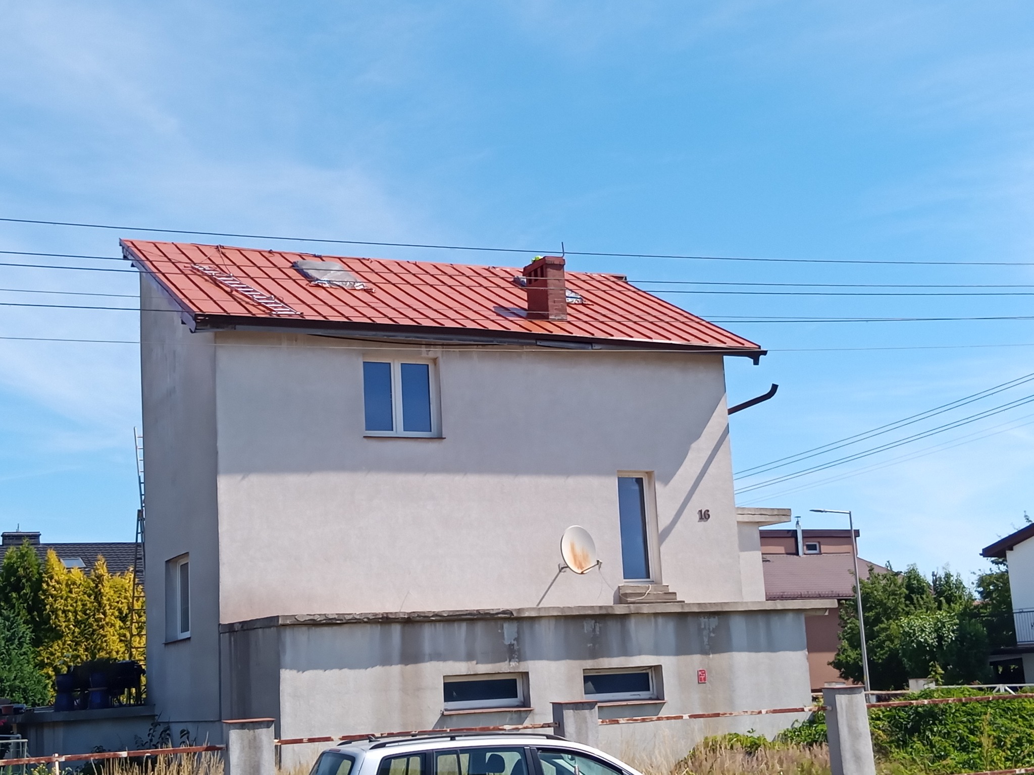 Dwupiętrowy dom z jasną elewacją i nowym, czerwonym dachem z blachodachówki, na którym leży drabina. Widoczne komin, okna dachowe i antena satelitarna.