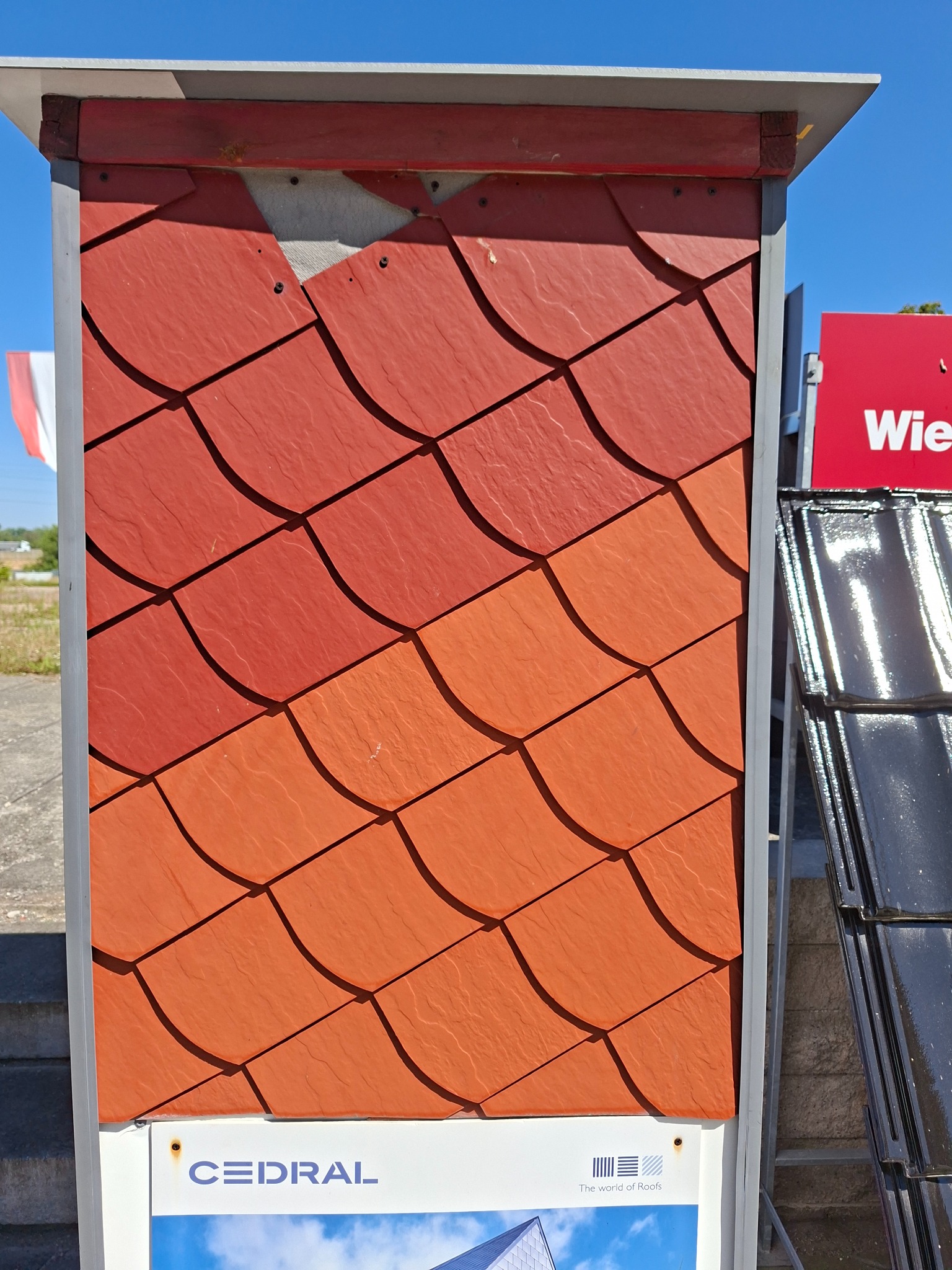 Panel ekspozycyjny z łuskowymi dachówkami Cedral w odcieniach czerwieni i pomarańczu, prezentujący różne kolory i faktury materiału, na tle błękitnego nieba.