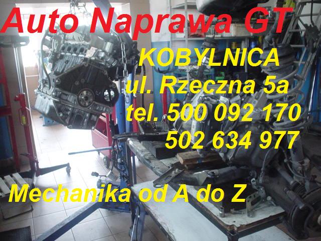 Silnik samochodowy zawieszony na łańcuchu w warsztacie, widoczne narzędzia i wyposażenie warsztatowe, w tle dane teleadresowe firmy.