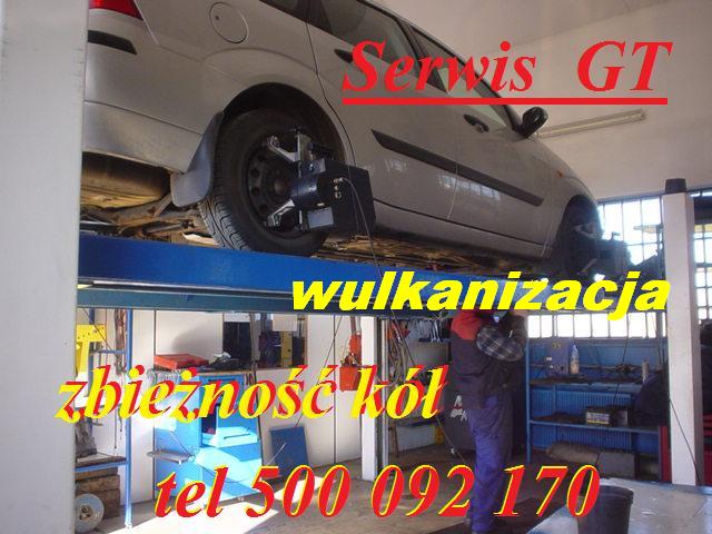 Samochód na podnośniku podczas ustawiania geometrii kół, widoczny pracownik serwisu i urządzenie do pomiaru zbieżności. W tle warsztat z wyposażeniem i napisy reklamowe na zdjęciu.