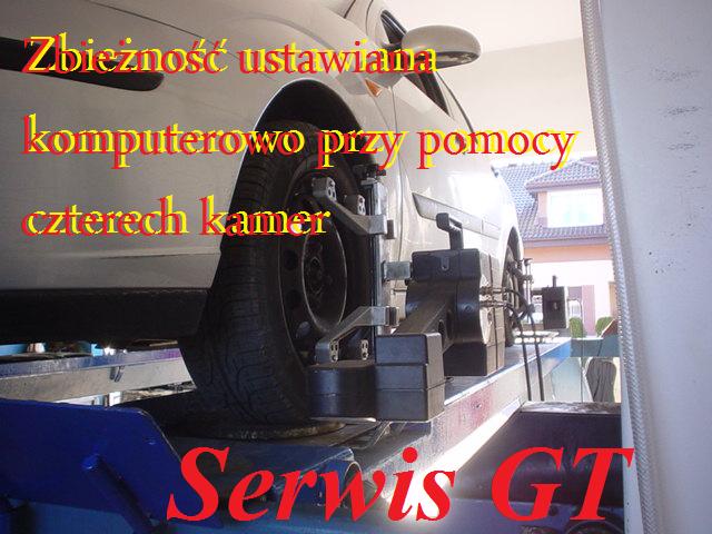 Ustawianie geometrii kół białego samochodu na podnośniku za pomocą systemu czterech kamer, widoczny fragment karoserii i koło z zamocowanym urządzeniem pomiarowym, w tle budynek mieszkalny.