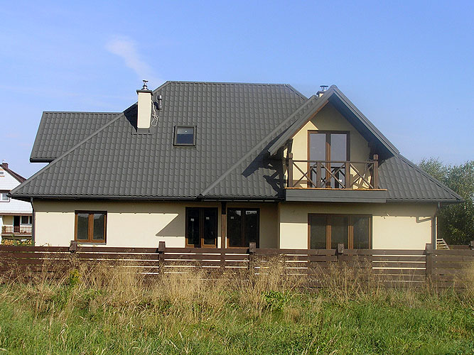 Gotowy dom z beżową elewacją i ciemnoszarą dachówką, widoczny balkon z drewnianą balustradą, ogrodzony niskim, drewnianym płotem. W tle błękitne niebo.